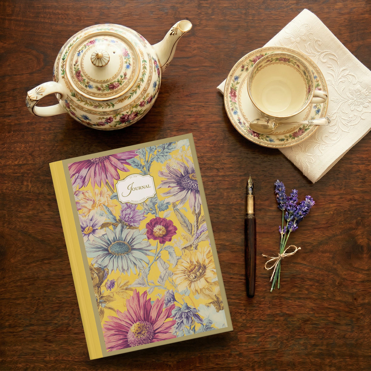 Tea set with teapot, cup, and saucer on a wooden table with a decorative journal, fountain pen, and lavender sprig.